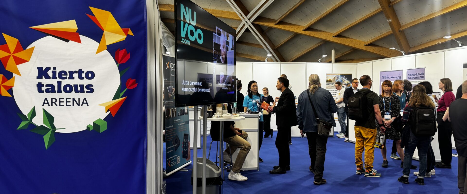 The event takes place in Oulu Hall. People are talking at the company exbihition tables. Floor is blue and the ceiling is brown and grey. At the left there is Circular Arena Oulu logo in a large blue banner. the TV next to it shows logo of Nuvoo.