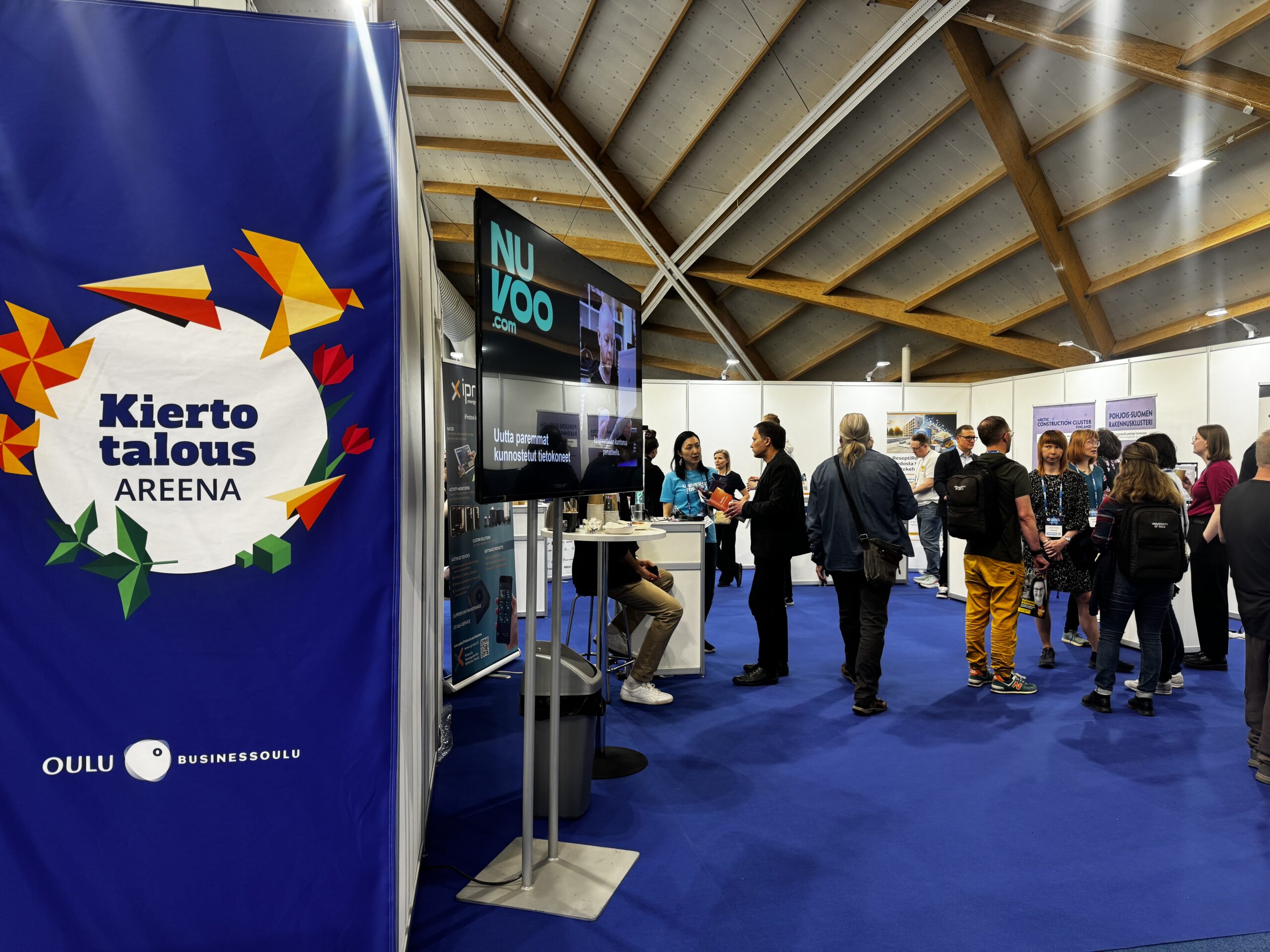 The event takes place in Oulu Hall. People are talking at the company exbihition tables. Floor is blue and the ceiling is brown and grey. At the left there is Circular Arena Oulu logo in a large blue banner. the TV next to it shows logo of Nuvoo.