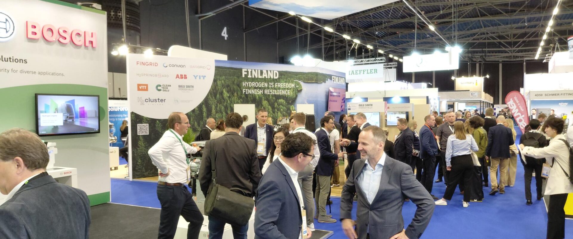 People are talking to each other in a convention centre. Floor has blue carpets and there is bright lights. Behind the people you can see wall text "Finland, Hydrogen is freedom, Finnish recilience."
