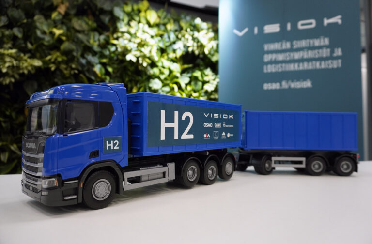 Blue toy truck with H2-letters. Behind it is some greenery.