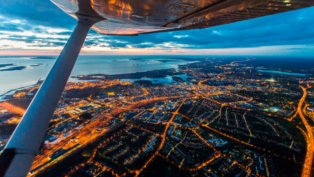 Lentokoneen siiven alta näkyy Oulu iltaruskossa, korkealta ilmasta käsin. Katuvalot loistavat keltaisena.