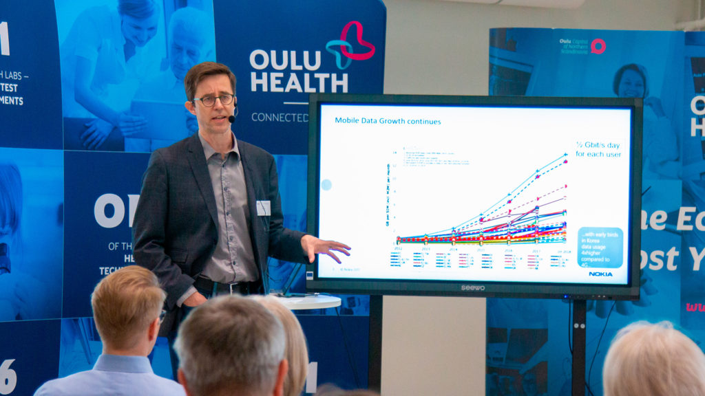 Man speaking to audience seen in the foreground. He has a large screen with the title ”Mobile Data Growth continues”. OuluHealth logo is shown in the background.