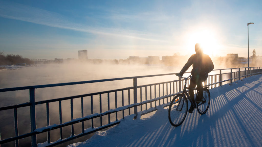 A person is biking on a winter’s day on the dam bridge. The person has dark clothes on, and sun is shining behind them. In the background, vapour is rising from the river, and buildings along the Oulu River can be seen on the bank.