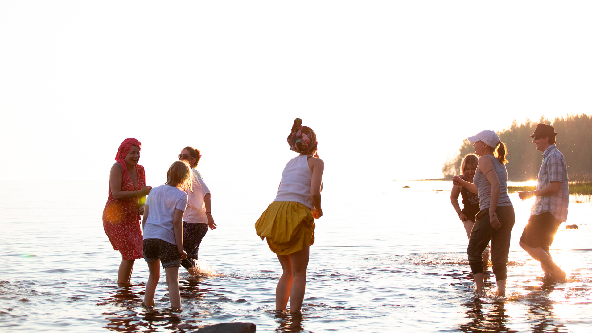 Nauravia ihmisiä kahlaamassa rantavedessä laskevan auringon kirkkaassa paisteessa. Kahlaajat ovat käärineet lahkeensa tai nostaneet hameenhelmojaan.
