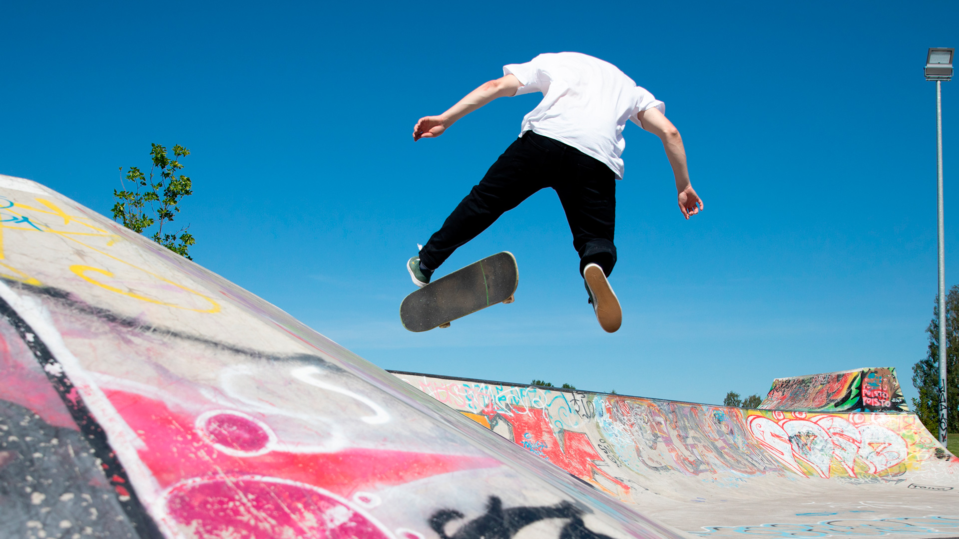 A person wearing a white shirt and black pants is skateboarding in a skate park decorated with colourful graffiti underneath a blue sky. The person is in the middle of a trick, and the skateboard is rolling around under their feet.
