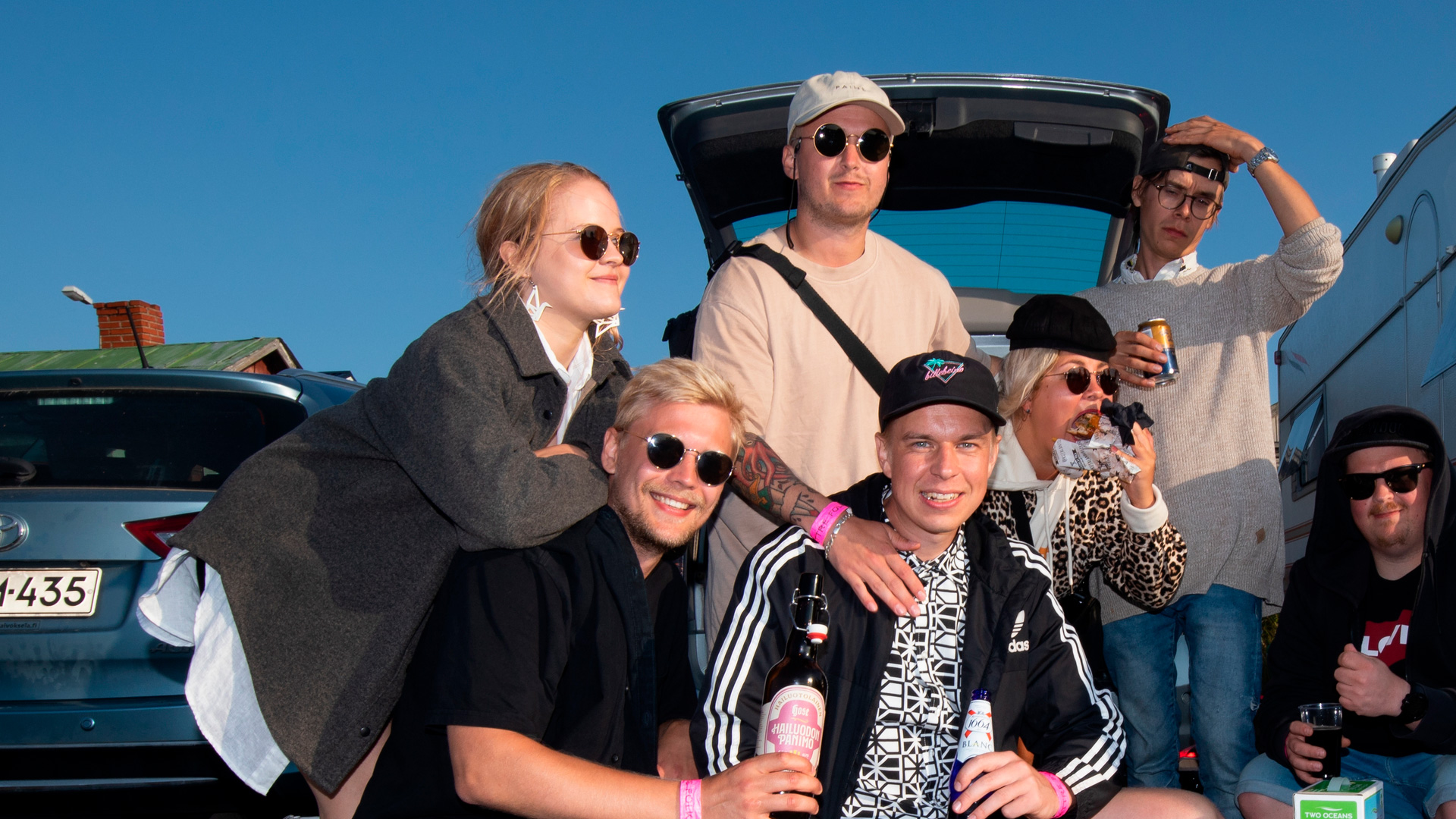 Young people are posing in front of the open trunk of a car. They are holding beverages and wearing relaxed clothes, some have sunglasses on.