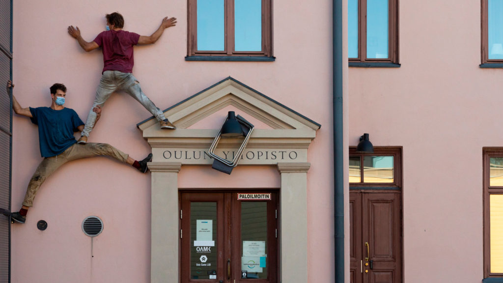 Two men have climbed up and are standing against the facade of a pink building. They are clinging to each other and parts of the facade for support. A text over a doorway on the facade says “Oulun yliopisto” (University of Oulu).