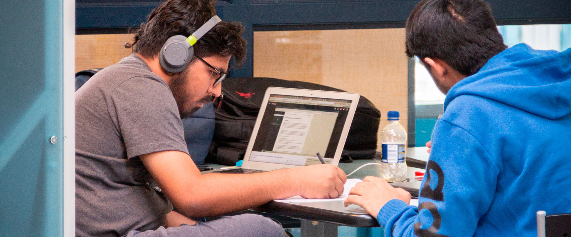 Two people are making notes in a room with glass walls. One is wearing headphones and has a laptop in front of them.