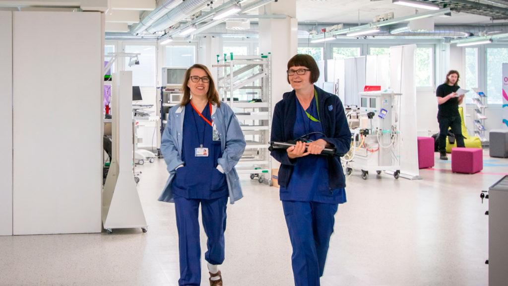 Two people wearing hospital employee outfits are smiling and walking toward the camera in a white room with hospital equipment showing in the background.