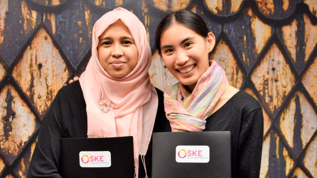 Two smiling women are standing in front of a wall with a pattern. One of the women has a scarf on her head, and they are both holding folders with the word “Oske” printed on them.
