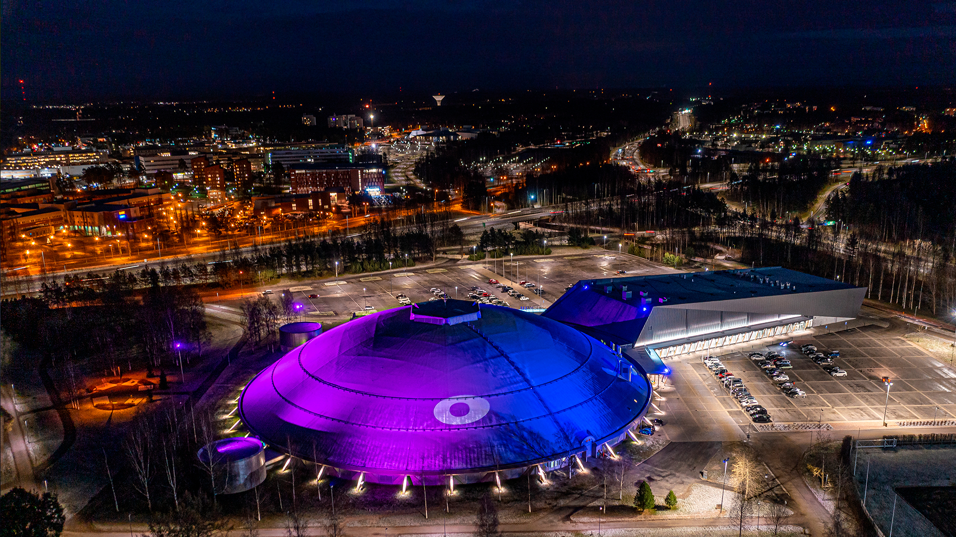 Ouluhalli arena and its surroundings photographed from the air. The roof of Ouluhalli has been coloured blue and violet with lights, and the degree sign of Oulu brand has also been created on the roof with lights.