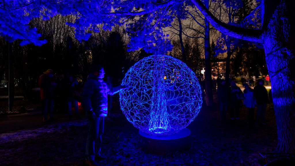 A spherical light art installation made from glowing fibres lights up its surroundings with blue colour in a park. There are people around it, and a person in the foreground is touching the installation.