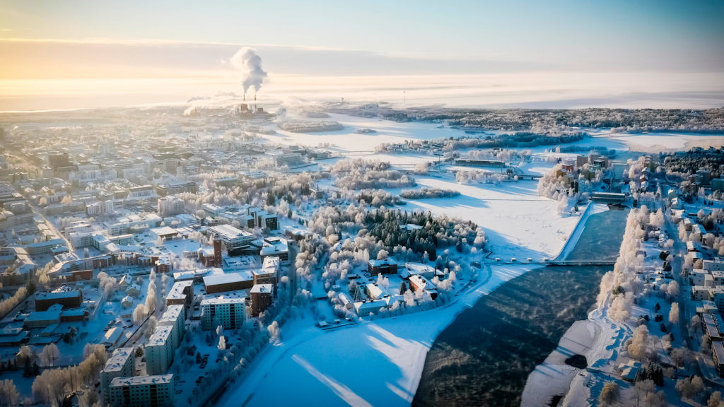 Talvinen Oulu kuvattuna ilmasta, Aurinko valaisee lumista kaupunkia kuvan vasemmasta reunasta, ja Oulujoki oikeassa reunassa virtaa sulana.