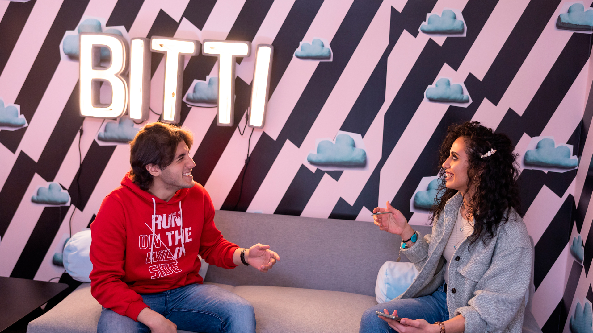 Two people are sitting on a sofa and laughing in front of a colourful wall. The wall has black and pink stripes and the shapes of clouds on it. One of the people has the word “bitti” written with light letters on the wall behind her.