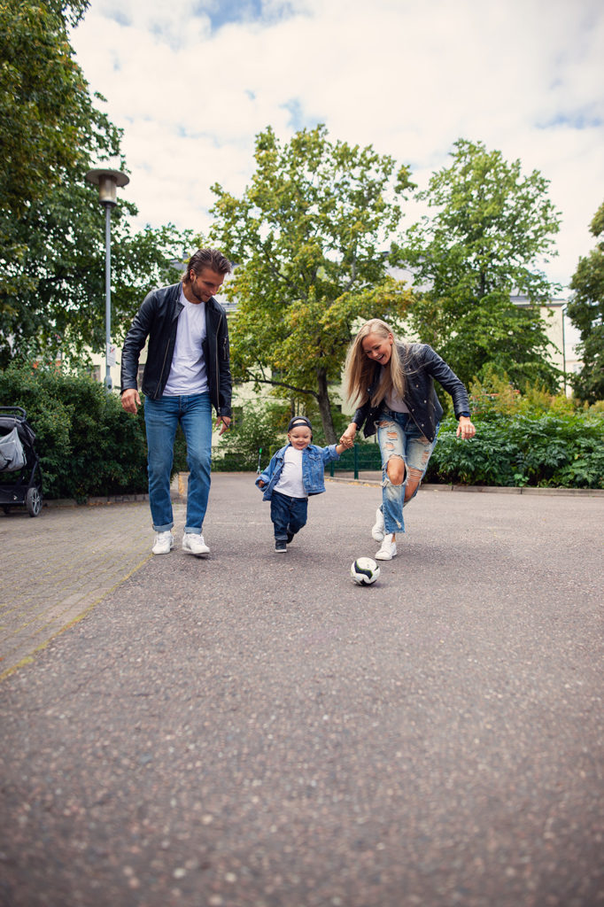 Vehreiden puiden keskellä isä, lapsi ja äiti potkivat pientä jalkapalloa iloisesti hymyillen.