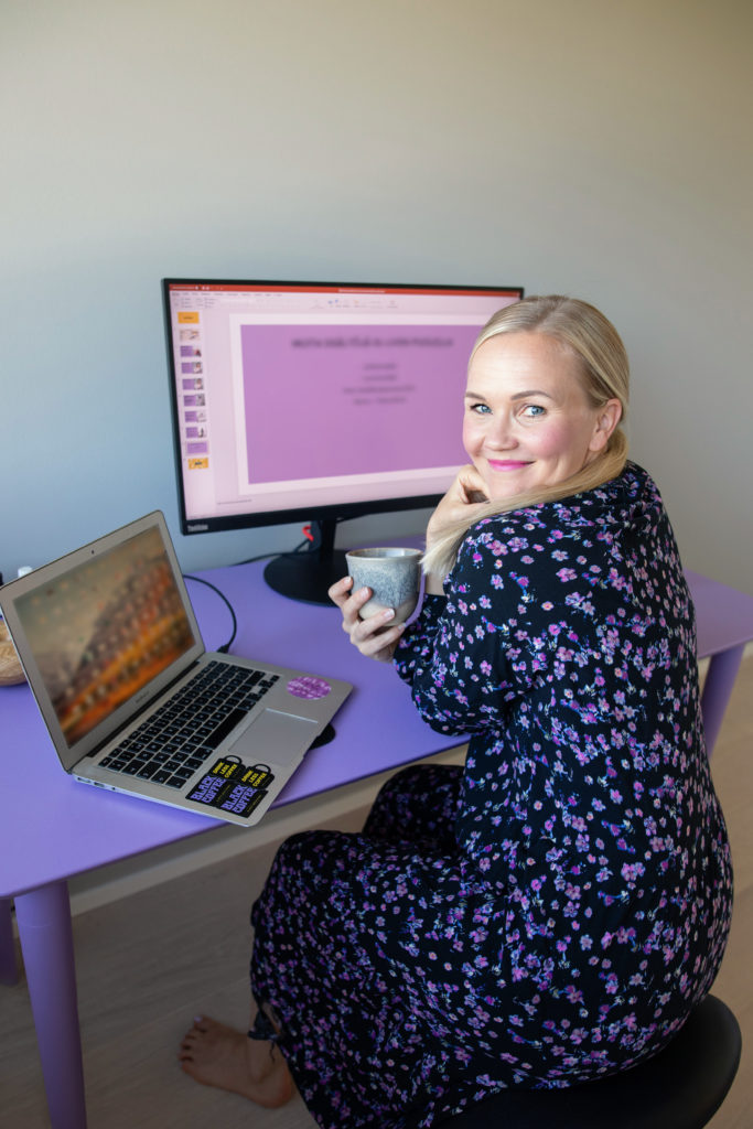 Henna istuu tietokoneen edessä ja hymyilee kameralle kahvikuppi kädellään. Hänellä on kukallinen mekko ja vaaleat hiukset.