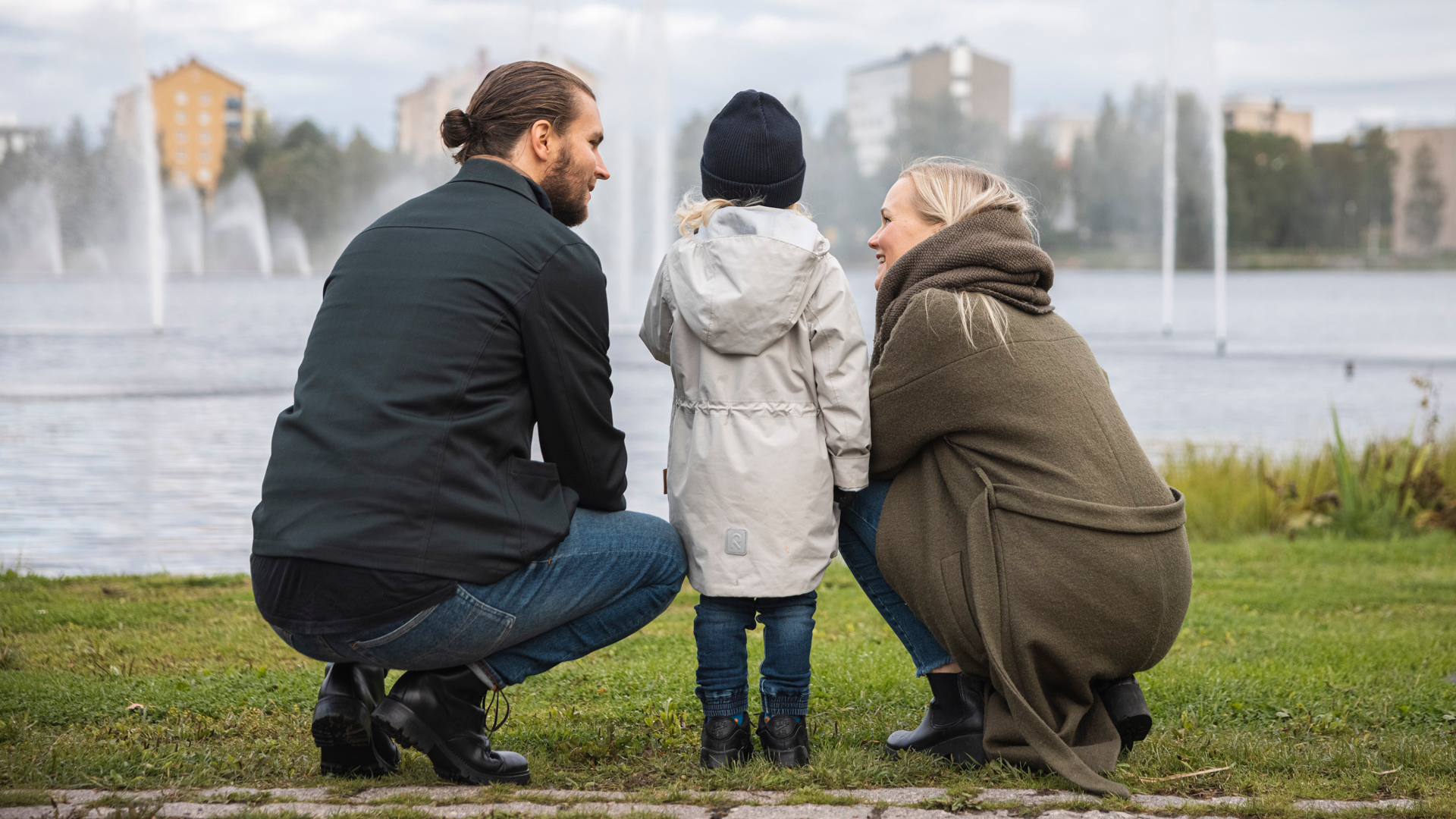 Isä, lapsi ja äiti selkä kameraan päin katsomassa Ainolanpuiston suhikulähteitä. Taustalla Toivoniemi.