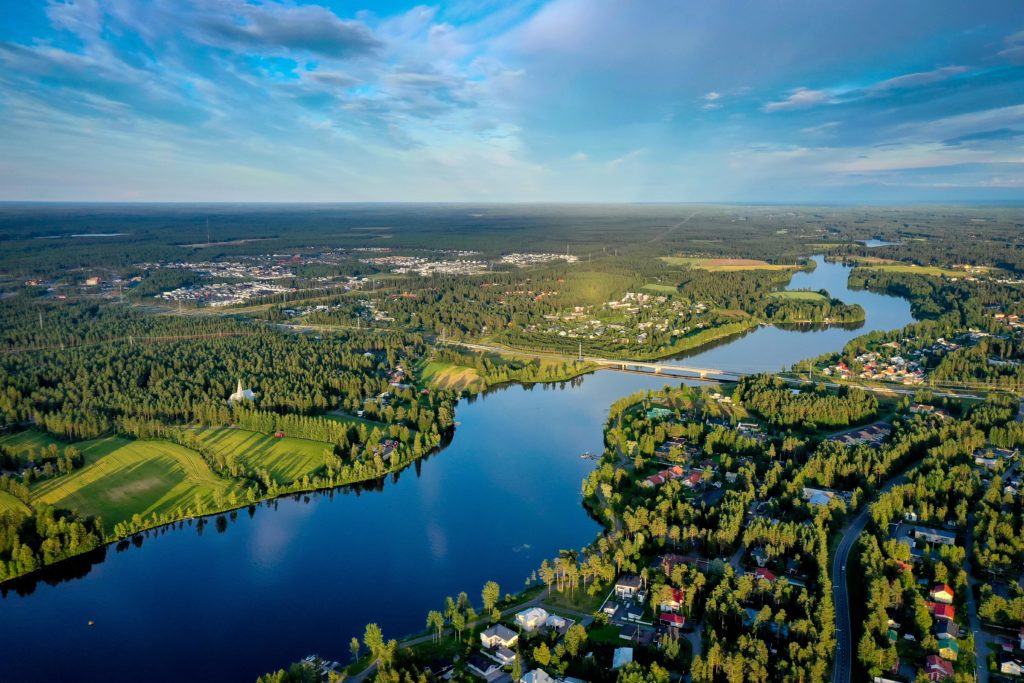 Aerial photo of the Oulu River and residences along the river, with trees and grass around them. The sky is blue with some clouds.