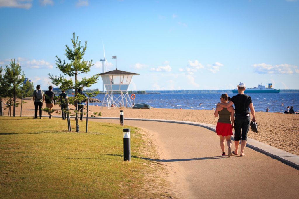Photograph of the Nallikari Beach. Two people are walking on a paved road in the foreground – one has their arm around the other one’s shoulders. The sky is blue and nearly cloudless. A ship is sailing on the sea.