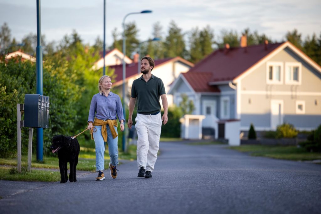 A woman and a man are walking a dog in a residential area with detached house on a summer evening. The dog is black and the woman has a coat tied around her waist.