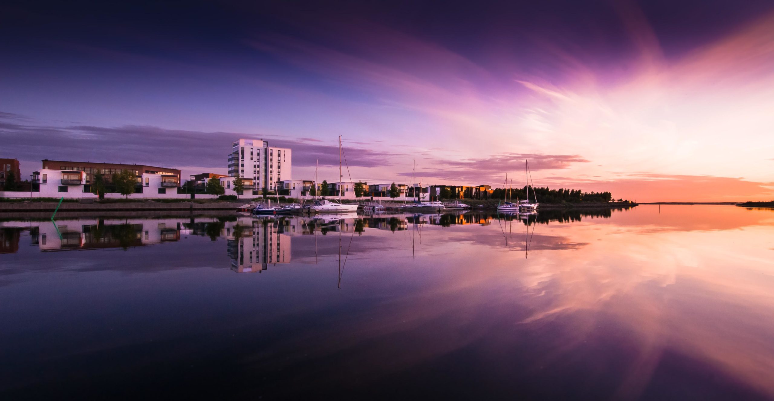 An evening dusk photographed by the water, colouring the sky with different shades of pink and yellow. On the opposite shore, there are tall apartment buildings and sailboats.