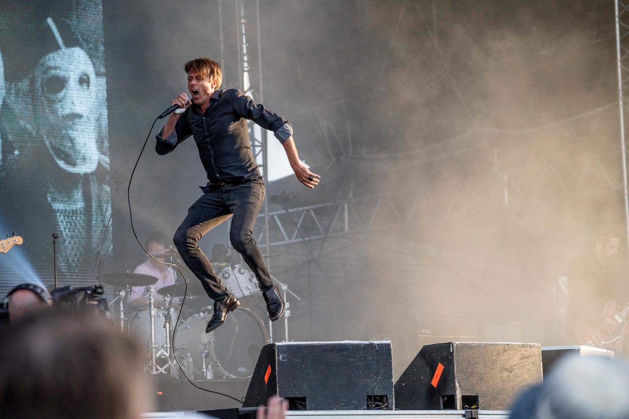 A man wearing black jeans on a stage jumps higher than his the monitors, holding a microphone.