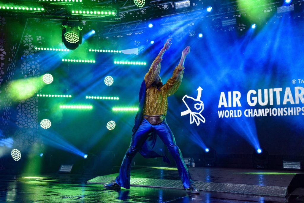 A person in the green and blue lights of a stage is lifting his hands in the air, wearing leather pants and a golden shirt. In the background is the text Air Guitar World Championships.	