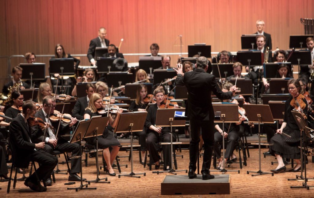 A symphony orchestra is rehearsing with a conductor who has his back to the camera, In the front row are string instruments and horns in the background. Everyone is wearing dark clothes.