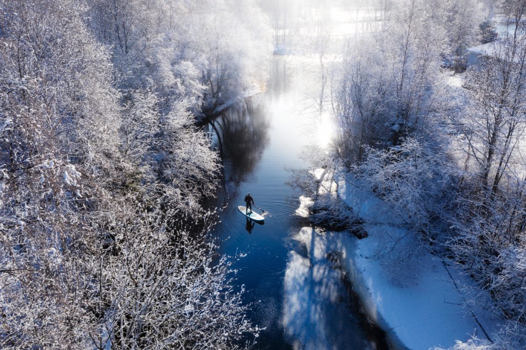 A person with an SUP board in a wintry landscape, photographed from above. The water is partly frozen and the trees are frosty.