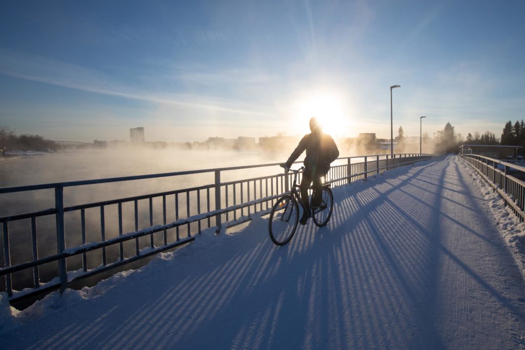 A person riding a bike on the Oulu Dam Bridge in the winter. Vapour is rising from the water and the rising sun is shining behind the cyclist.