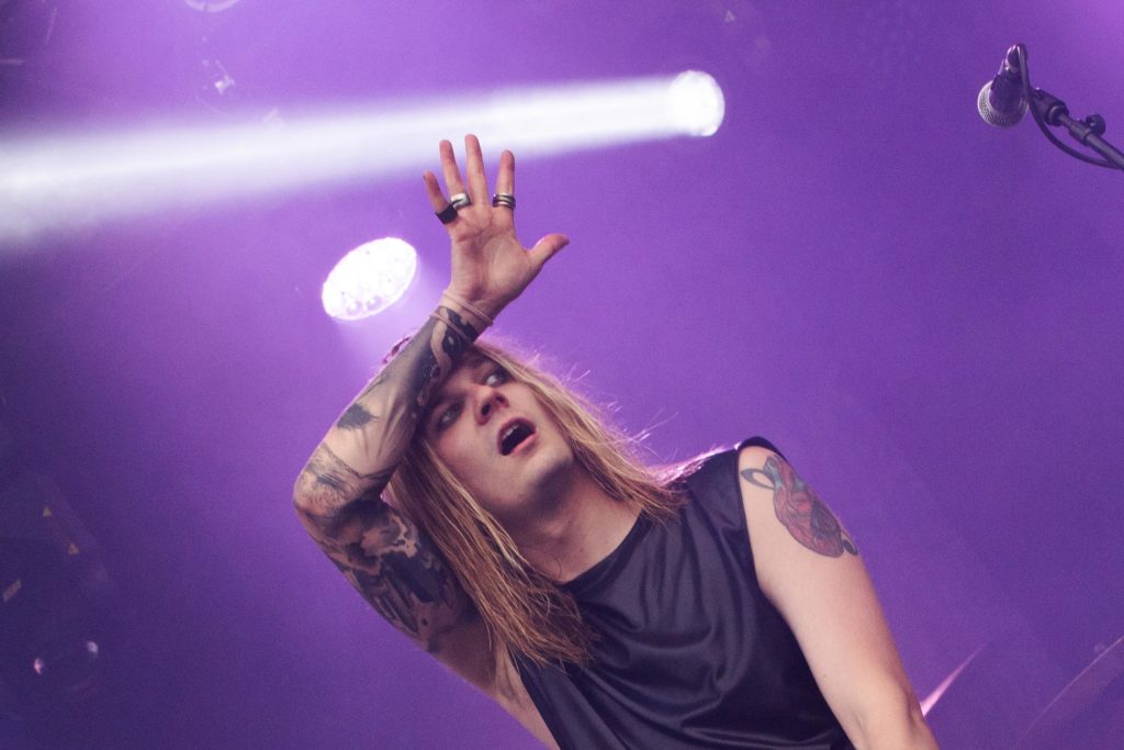 A long-haired, tattooed man has his arm on his forehead on a stage. The photo is slightly tilted, the background is purple and there is a microphone at the edge of the picture.
