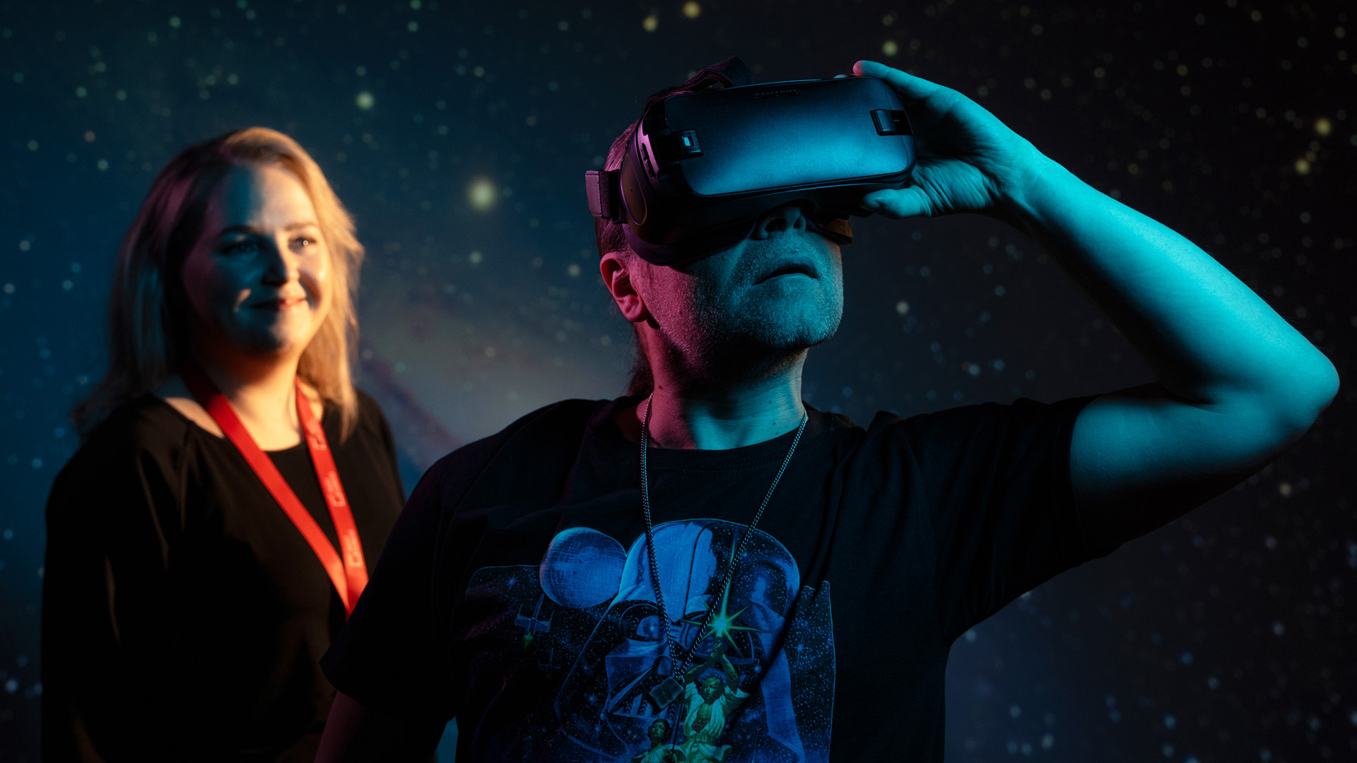 A woman and a man standing in front of a glittering wall. A man stares into the distance with VR glasses.