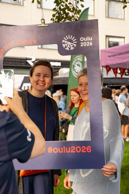 Kaksi henkilöä poseeraa Kesäillan kattaus -tapahtumassa iloisena Oulu2026-kehyksen kanssa, kun kolmas henkilö ottaa heistä valokuvaa.